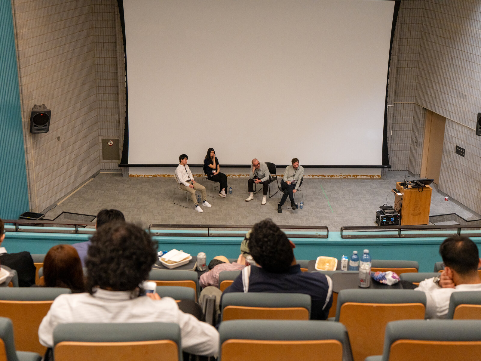 Four panelists on stage with a podium and sound equipment, audience in foreground.