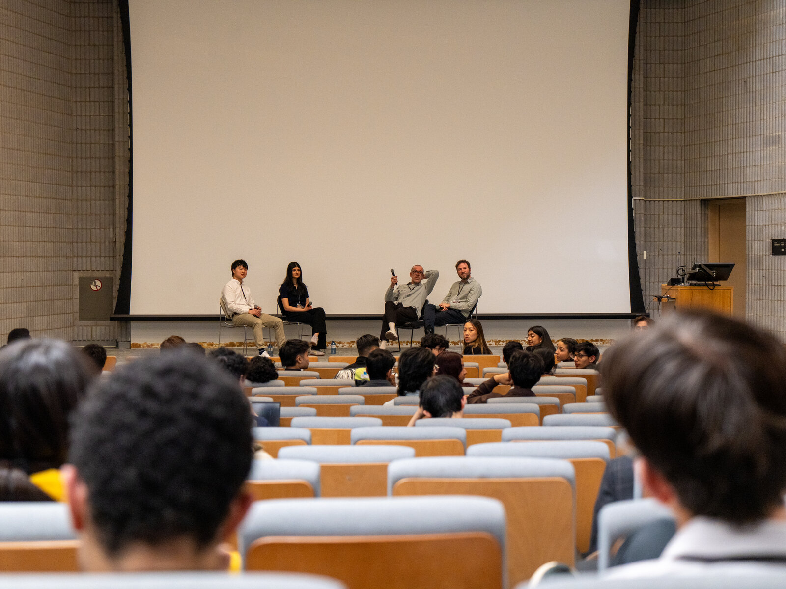 Four panelists on stage with a No Smoking sign, seen from the audience in a lecture hall.